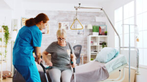 Woman seated on edge of hospital bed, poised to stand up, while another woman in hospital scrubs is standing next to her, helping her to position forearm crutches.