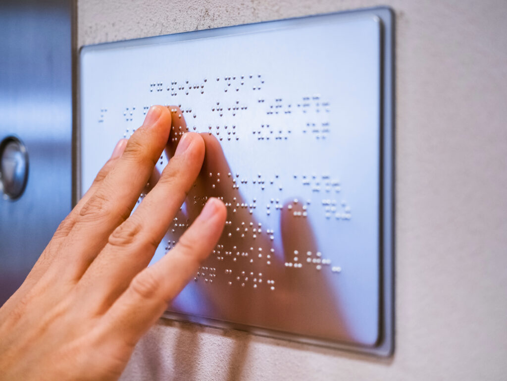 Hand reading a Braille sign mounted on a wall.