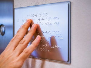 Hand reading a Braille sign mounted on a wall.