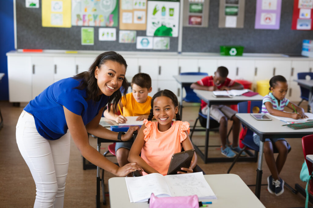 Teacher assisting a student using a tablet while other students work independently in a classroom.