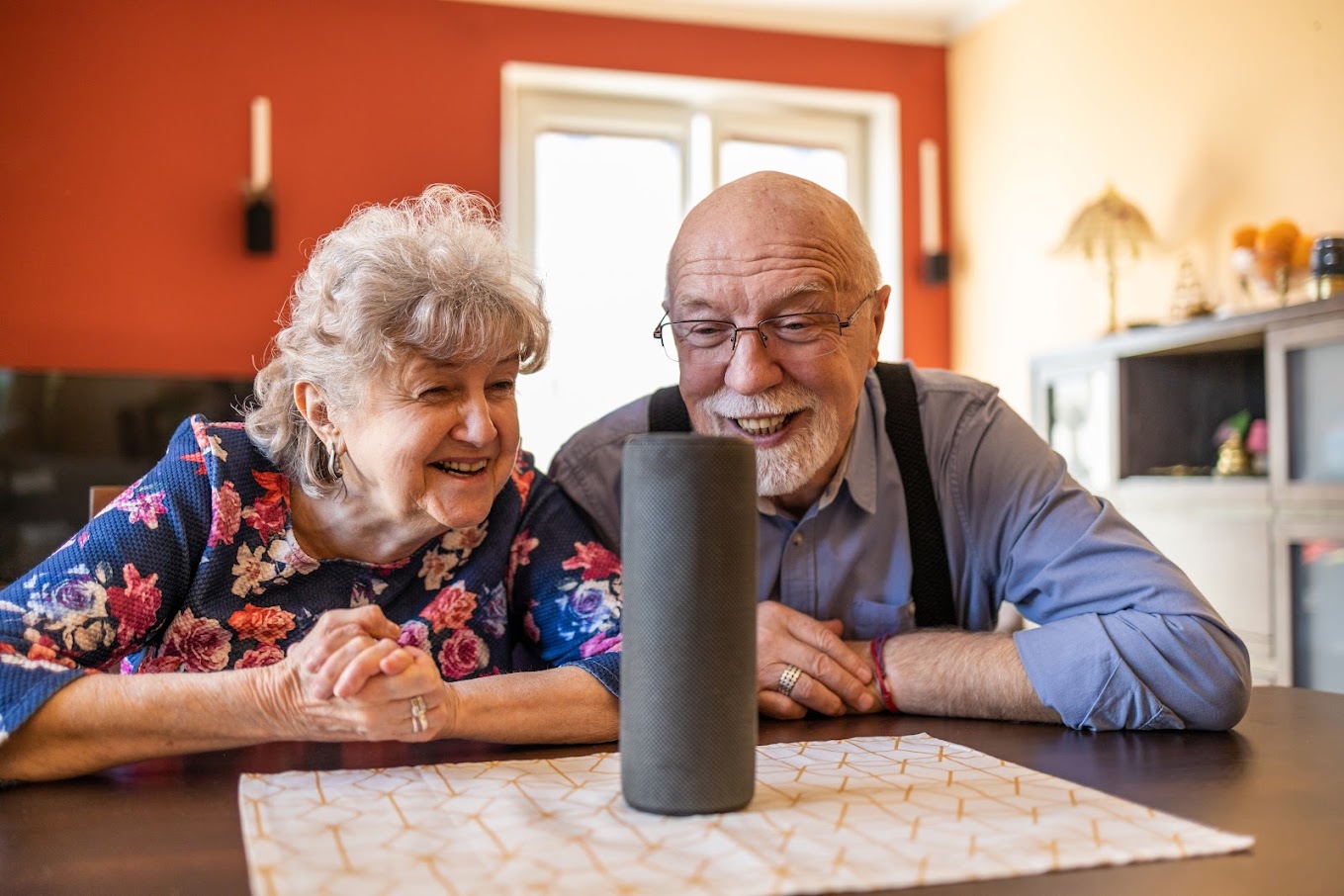 An older couple smiling and sitting at a table with a smart speaker in front of them on the table